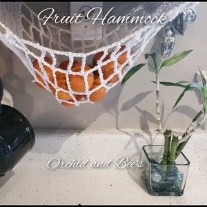 May include: A white crocheted fruit hammock hanging from a cabinet with oranges inside. The hammock is labelled "Fruit Hammock". A potted bamboo plant is on the counter below the hammock. The counter is labelled "Orchid and Bee's".