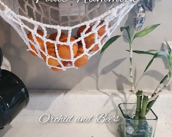 May include: A white crocheted fruit hammock hanging from a cabinet with oranges inside. The hammock is labelled "Fruit Hammock". A potted bamboo plant is on the counter below the hammock. The counter is labelled "Orchid and Bee's".