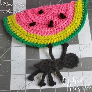May include: A crocheted watermelon with a green and yellow rind and a pink center. A black crocheted ant is hanging from the watermelon. The text "Watermelon Ant Thief" is written above the watermelon.