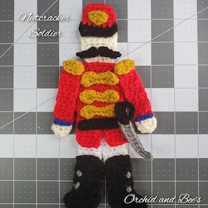 May include: A handmade crochet nutcracker soldier in red, black, white, and gold. The soldier has a mustache, a hat, and a sword. The text "Nutcracker Soldier" and "Orchid and Bee's" are visible.