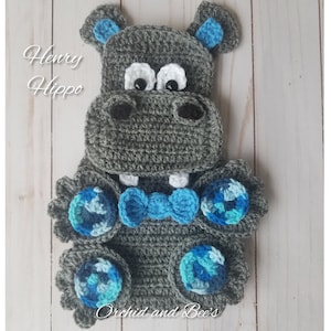 May include: A gray crocheted hippopotamus with blue and white accents. The hippopotamus is wearing a blue bow tie and has a white crocheted nose. The hippopotamus is sitting on a white wooden surface. The text "Henry Hippo" is written in black letters on the surface.