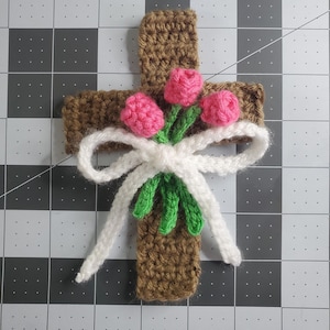 May include: A brown crocheted cross with a white bow and pink flowers.