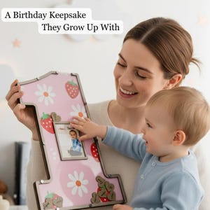 Puede incluir: Un marco de fotos rosa con forma de n&uacute;mero uno con un estampado de fresas y margaritas. El marco contiene una foto peque&ntilde;a y adornos de madera en forma de coraz&oacute;n. El texto en el marco dice: "A Birthday Keepsake They Grow Up With."