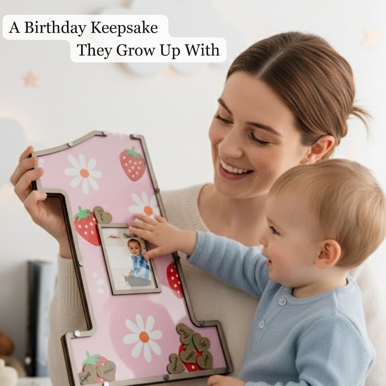 Puede incluir: Un marco de fotos rosa con forma de n&uacute;mero uno con un estampado de fresas y margaritas. El marco contiene una foto peque&ntilde;a y adornos de madera en forma de coraz&oacute;n. El texto en el marco dice: "A Birthday Keepsake They Grow Up With."