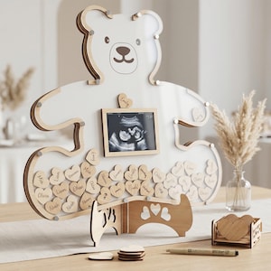 May include: A white bear-shaped guest book with a wooden frame holding an ultrasound image. Wooden heart-shaped name tags are included. The bear has a light brown outline and a small wooden stand.