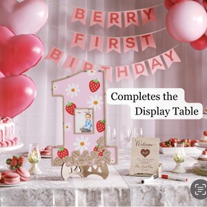 Puede incluir: Un letrero rosa con el n&uacute;mero uno con un patr&oacute;n de fresas y margaritas, con una foto. El letrero es parte de una exhibici&oacute;n de primer cumplea&ntilde;os, con una pancarta que dice "Berry First Birthday".