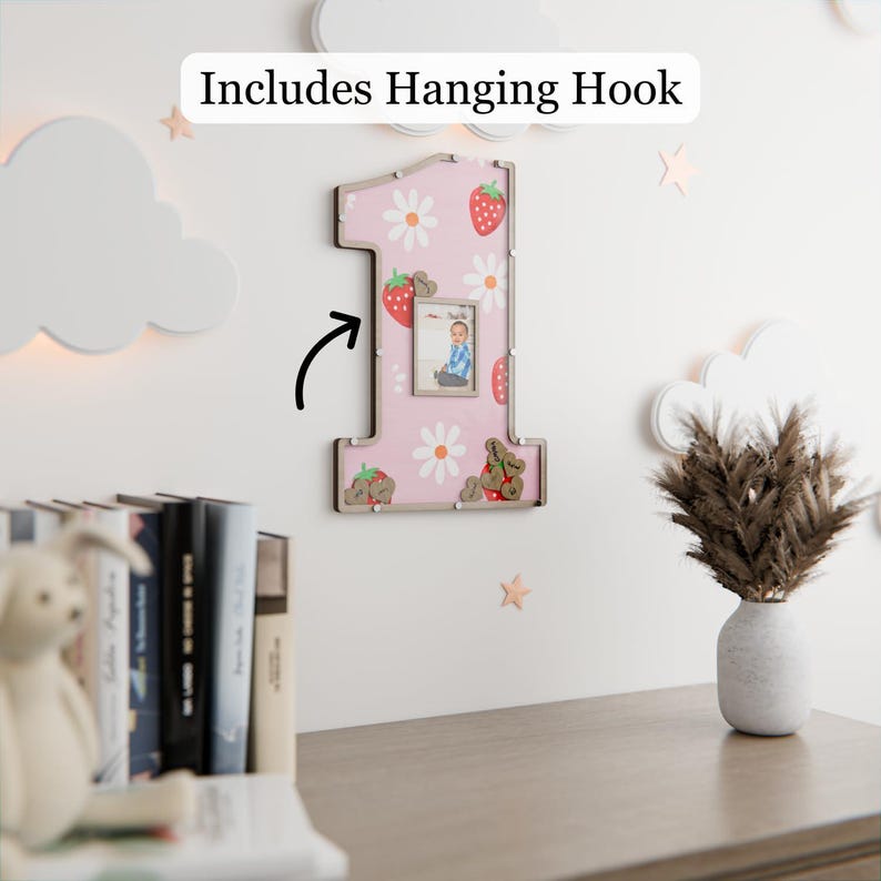 Puede incluir: Un marco de fotos rosa con forma de n&uacute;mero uno, decorado con fresas y margaritas. El marco incluye una foto peque&ntilde;a y est&aacute; decorado con fresas y osos. El texto "Includes Hanging Hook" es visible en la parte superior.