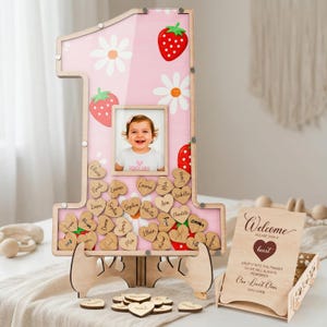 Puede incluir: Un marco de madera rosa con el n&uacute;mero uno, con una foto de beb&eacute;, fresas y margaritas. Incluye etiquetas con nombres en forma de coraz&oacute;n de madera y un letrero de bienvenida. El marco est&aacute; sostenido por un soporte de madera. Una alternativa &uacute;nica al libro de visitas.