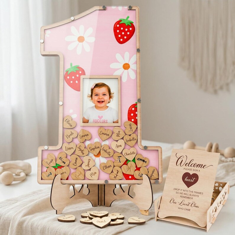 Puede incluir: Un marco de madera rosa con el n&uacute;mero uno, con una foto de beb&eacute;, fresas y margaritas. Incluye etiquetas con nombres en forma de coraz&oacute;n de madera y un letrero de bienvenida. El marco est&aacute; sostenido por un soporte de madera. Una alternativa &uacute;nica al libro de visitas.