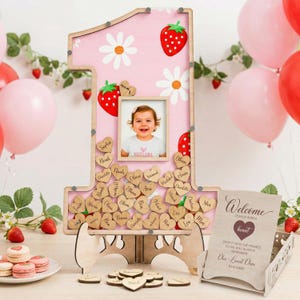 Puede incluir: Un libro de visitas de madera rosa con forma de n&uacute;mero uno con un marco de fotos, dise&ntilde;os de fresas y margaritas. Incluye etiquetas de madera en forma de coraz&oacute;n con nombres y un letrero de bienvenida. Foto de una ni&ntilde;a en el marco.