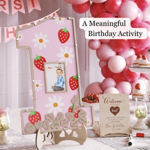 Puede incluir: Un letrero de cumplea&ntilde;os rosa con forma de n&uacute;mero uno con un marco de fotos, fresas y margaritas. El letrero tiene una base de madera e incluye etiquetas con nombres en forma de coraz&oacute;n. El texto en el letrero dice "A Meaningful Birthday Activity."