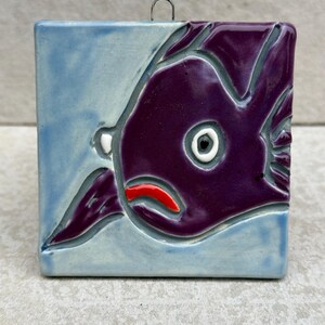 May include: A purple ceramic tile with a stylized fish design. The fish has a white eye, a red mouth, and a blue background.