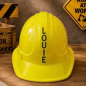 May include: A bright yellow hard hat featuring the name LOUIE printed in bold, dark green capital letters vertically down the centre. The protective construction helmet has a smooth, rounded surface and a wide brim, designed for safety gear.