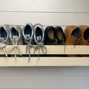 May include: A wooden shoe rack with two shelves, holding six pairs of shoes. The top shelf has three pairs of grey trainers and two pairs of black dress shoes. The bottom shelf has one pair of brown leather shoes.