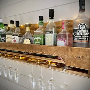 Może przedstawiać: Ruska drewniana p&oacute;łka ścienna z wbudowanym stojakiem na kieliszki do wina. P&oacute;łka jest wypełniona r&oacute;żnymi butelkami alkoholu, w tym Jack Daniels, Jim Beam i Russian Standard Vodka. P&oacute;łka jest oświetlona lampkami choinkowymi.