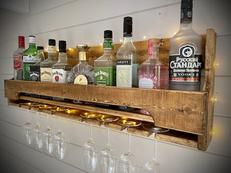 Może przedstawiać: Ruska drewniana p&oacute;łka ścienna z wbudowanym stojakiem na kieliszki do wina. P&oacute;łka jest wypełniona r&oacute;żnymi butelkami alkoholu, w tym Jack Daniels, Jim Beam i Russian Standard Vodka. P&oacute;łka jest oświetlona lampkami choinkowymi.