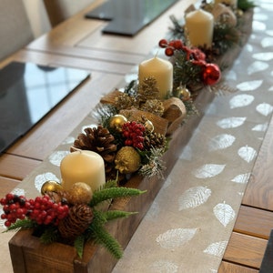 May include: A wooden box centrepiece with candles, pine cones, greenery, red berries, and gold ornaments. The centrepiece is on a table runner with a silver leaf pattern.