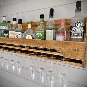 Może przedstawiać: Drewniana, wisząca p&oacute;łka na alkohol z g&oacute;rną p&oacute;łką na butelki i dolną p&oacute;łką na kieliszki do wina. P&oacute;łka jest wykończona ciemnobrązowym lakierem i ma rustykalny wygląd. P&oacute;łka jest wypełniona r&oacute;żnymi butelkami alkoholu, w tym w&oacute;dką, dżinem, rumem i whisky. P&oacute;łka ma r&oacute;wnież rząd kieliszk&oacute;w do wina wiszących na dole.