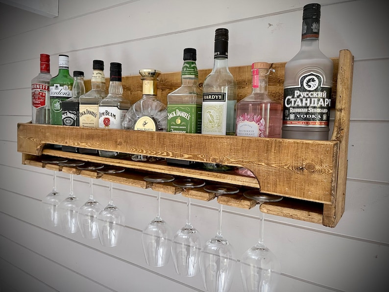 Może przedstawiać: Drewniana, wisząca p&oacute;łka na alkohol z g&oacute;rną p&oacute;łką na butelki i dolną p&oacute;łką na kieliszki do wina. P&oacute;łka jest wykończona ciemnobrązowym lakierem i ma rustykalny wygląd. P&oacute;łka jest wypełniona r&oacute;żnymi butelkami alkoholu, w tym w&oacute;dką, dżinem, rumem i whisky. P&oacute;łka ma r&oacute;wnież rząd kieliszk&oacute;w do wina wiszących na dole.