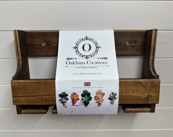 Puede incluir: Una estantería de pared de madera rústica con dos compartimentos. La estantería está teñida de un color marrón oscuro y tiene un acabado desgastado. La estantería está etiquetada con una tarjeta blanca que dice "Oakham Creations Fine Artisan Creations www.oakham-creations.co.uk Proudly designed and manufactured in Great Britain".