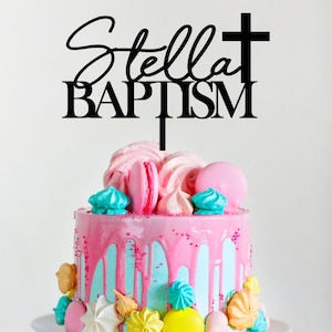 Puede incluir: Topper de pastel de acrílico negro con el texto "Stella Baptism" y una cruz. El topper está en un pastel rosa y azul decorado con macarons y merengues.