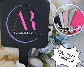 Puede incluir: Espejo de mano negro con un diseño circular que presenta las letras "AR" en rosa y el texto "Beauty & Lashes" en blanco. El espejo tiene una superficie de espejo real y está disponible en varios colores.