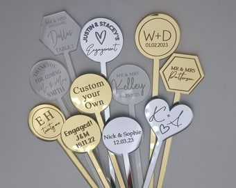 May include: A collection of personalized drink stirrers in gold and silver. Each stirrer features custom text, including names, dates, and phrases like "Engaged!" and "Engagement." The stirrers have various shapes, including circles, hearts, and hexagons.