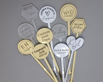 May include: Assortment of gold and silver acrylic drink stirrers with personalized text. The stirrers feature various shapes, including circles, hearts, and hexagons. The text includes names, dates, and messages such as "Engaged!" and "Custom your own".