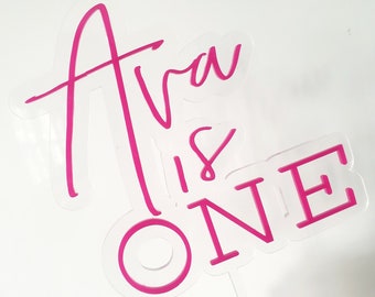 Puede incluir: Topper de pastel de acrílico rosa con el texto "Ava is One" en una fuente de letra moderna. El número uno está en negrita, en fuente de bloque.