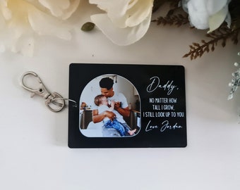 Puede incluir: Llavero rectangular negro con un mosquetón plateado. El llavero presenta una foto de un padre y un niño dentro de un marco arqueado blanco. El texto dice "Daddy, no matter how tall I grow, I still look up to you. Love Jordan."