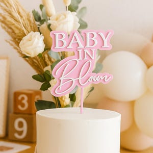 Może przedstawiać: Różowy i biały topper na ciasto z napisem "BABY IN BLOOM" umieszczony na wierzchu białego, lukrowanego ciasta. Ciasto jest udekorowane kompozycją kwiatową i drewnianymi klockami z cyframi 3 i 9. Balony w tle.