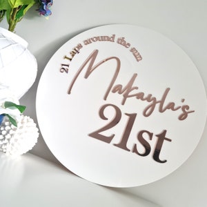 May include: White round sign with the text "21 Laps around the sun Makayla's 21st" in rose gold lettering.