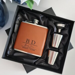 May include: A tan leather hip flask with the initials "BD" and the name "Barry Finn" engraved on the front. The flask is presented in a black gift box, alongside two stainless steel shot glasses and a funnel. The text "HUSBAND o FATHER" is below the name.