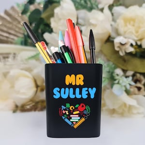 May include: Black pencil holder with the text "MR SULLEY" in yellow and blue lettering. A heart shape filled with colorful school supplies is below the text.
