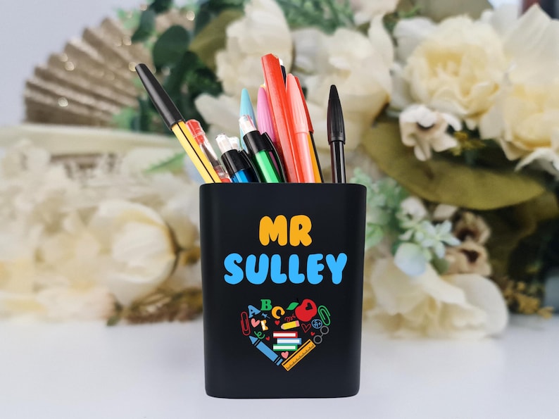 May include: Black pencil holder with the text "MR SULLEY" in yellow and blue lettering. A heart shape filled with colorful school supplies is below the text.