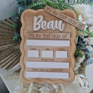 以下が含まれることがあります： 白い筆記体で「Beau」という名前が入った木製の看板。看板には「私の最後の日」と「私の最初の日」というフレーズが含まれています。日付、年齢、身長、好きなもの、将来の願望のためのスペースがあります。看板は花に囲まれています。