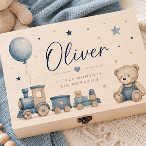 May include: A rectangular wooden keepsake box featuring a printed illustration of a blue train, a teddy bear, a balloon, and stars. The text reads Oliver, Little Moments, Big Memories. The box has a metal latch on the front edge for closure.