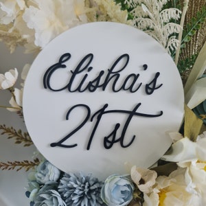 以下が含まれることがあります： 白い丸い看板に、黒い筆記体で「Elisha's 21st」と書かれています。看板は白と青の花に囲まれています。