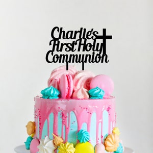May include: Black acrylic cake topper with the text "Charlie's First Holy Communion" and a cross. The topper is on a pink and blue cake decorated with macarons and meringue.