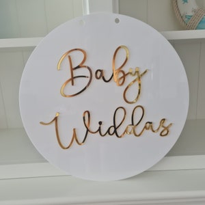 May include: White round acrylic sign with gold lettering that says "Baby Wildas".