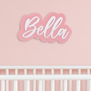 May include: A pink acrylic name sign with the name "Bella" in white script lettering. The sign is cloud-shaped and mounted on a pink wall, above a white cot.