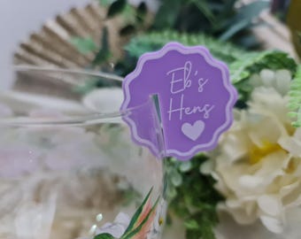 May include: Purple favor tag with the text "Eb's Hens" and a white heart. The tag is attached to a clear glass with a floral design.
