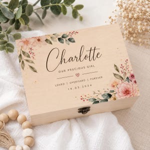 May include: A rectangular light-coloured wooden keepsake box featuring a metal latch. The lid is printed with floral illustrations in pink and green, the name Charlotte, the text OUR PRECIOUS GIRL, LOVED | CHERISHED | FOREVER, and 14.03.2024.