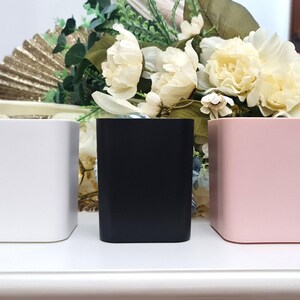 May include: Three square containers, one white, one black, and one pink, are arranged in a row on a white surface.