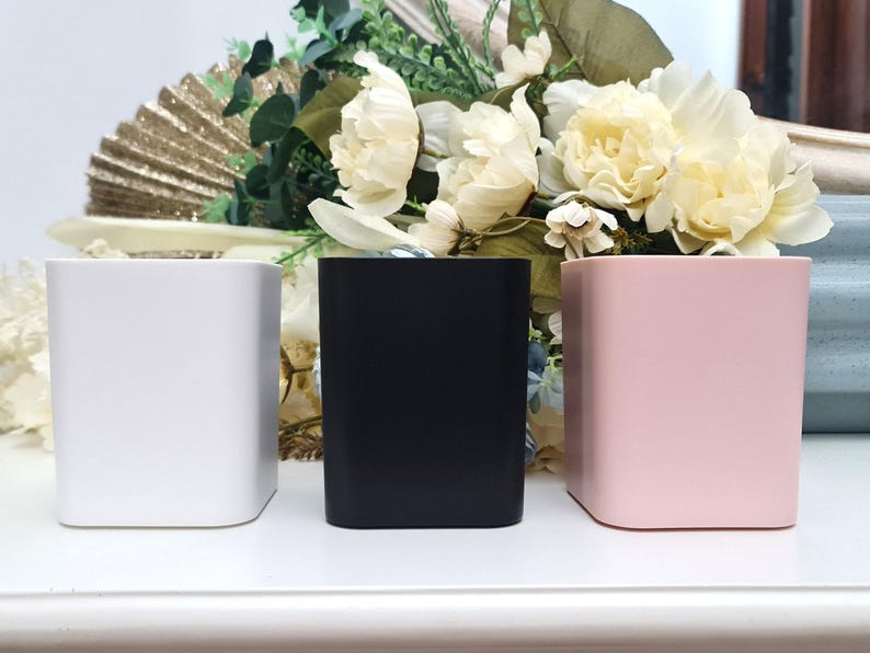 May include: Three square containers, one white, one black, and one pink, are arranged in a row on a white surface.