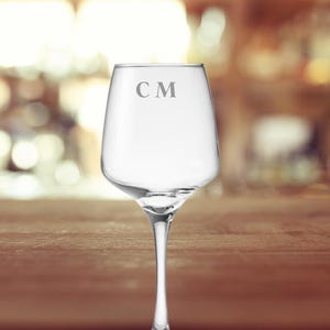 May include: A clear wine glass with a long stem, engraved with the initials "CM". The glass is empty and sits on a wooden surface. The background is blurred, suggesting a bar or restaurant setting.