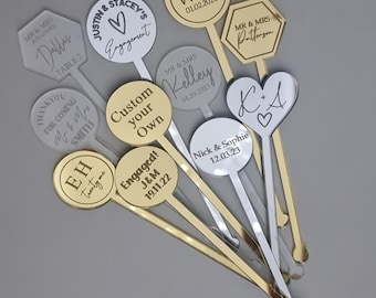 Puede incluir: Surtido de agitadores de bebidas de acrílico dorado y plateado con mensajes personalizados. Los agitadores presentan diversas formas, incluyendo círculos, corazones, hexágonos y un diamante. Los mensajes incluyen nombres, fechas y frases como "¡Compromiso!" y "Personaliza el tuyo".