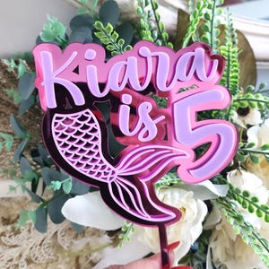 Op de afbeelding: Een roze en paarse acryl taarttopper met de tekst "Kiara is 5" en een zeemeerminstaartontwerp. De topper is ontworpen voor een vijfde verjaardag en wordt vastgehouden door een heldere stok.