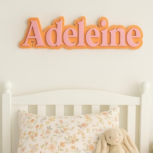 Peut inclure: Panneau de nom rose et orange avec le nom "Adeleine" dans une police fantaisie. Le panneau est fixé sur un mur crème au-dessus d'un cadre de lit blanc, d'un oreiller floral et d'un lapin en peluche.