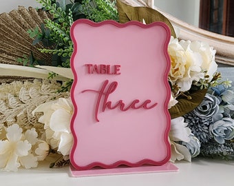 Puede incluir: Número de mesa rosa con borde ondulado. El letrero dice "TABLE Three" en rosa, en letra cursiva.